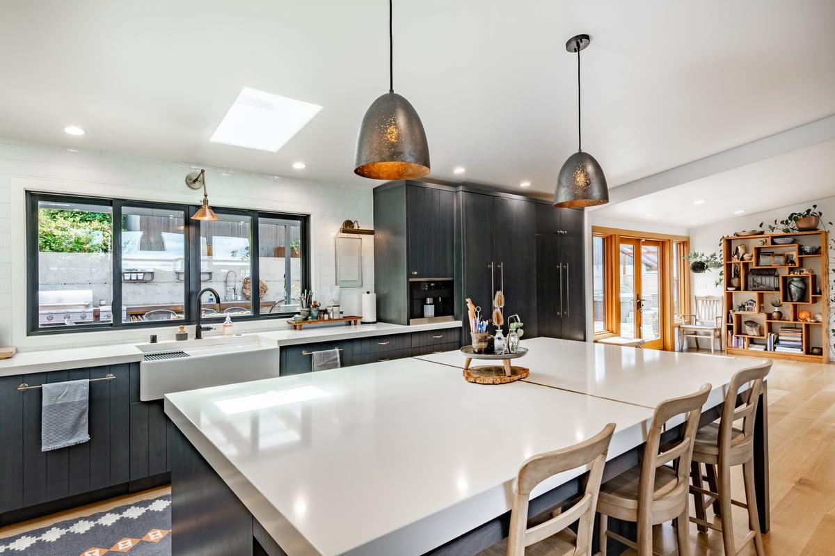 A modern kitchen with dark cabinetry light countertops warm wood flooring. Featuring a large central island with white countertop and seating for four using wooden stools.